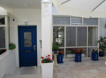 בכניסה לבית: דלת כחולה, חלון גדול, שורת עציצים עם גרניום...יוון זה כאן
