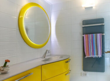 אמבטיית ילדים. ארון אמבטיה עם חזית זכוכית צהובה, ומחמם מגבות כחול על הקיר.