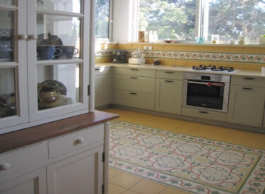 אחרי שיפוץ הדירה: עיצוב המטבח -- ארונות בצבע ירקרק, חלונות ענקיים, שטיח מאריחים מעוטרים בריצוף וחיפ קיר תואם לכך.