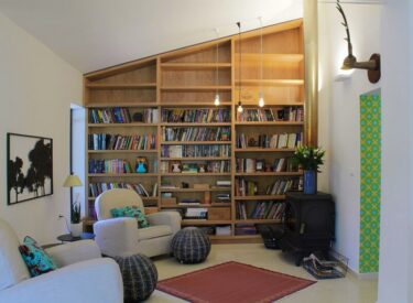 קיר ספרייה ואח לעצים מהווים עוגן לעיצוב חדר משפחה צבעוני וחמים. חדר זה פתוח לסלון, המעוצב בסגנון אלגנטי.